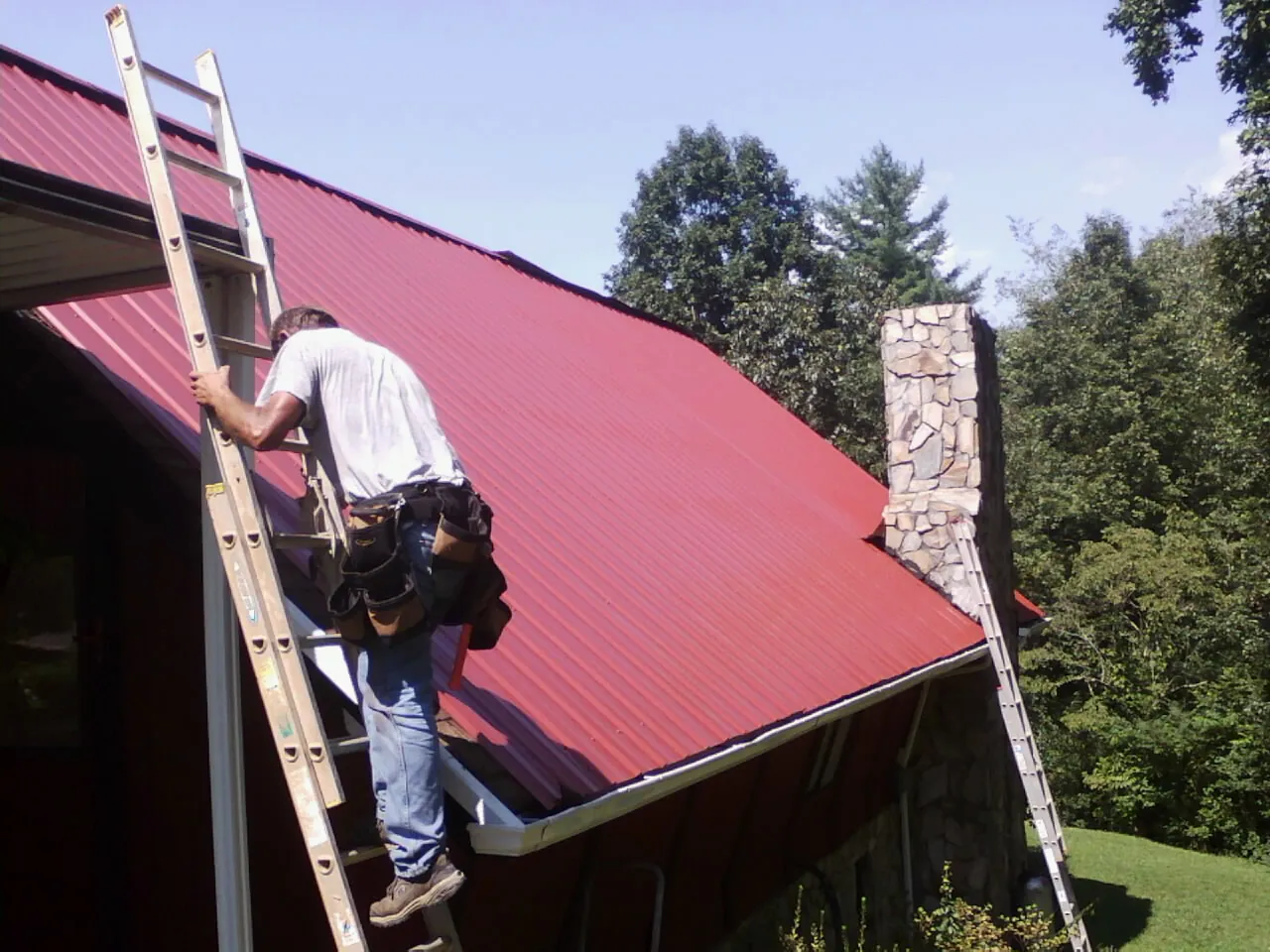 Roofer on a ladder beside a red metal roof and stone chimney — Soffit & Fascia Repair by Roofing Brothers in Snow Hill
